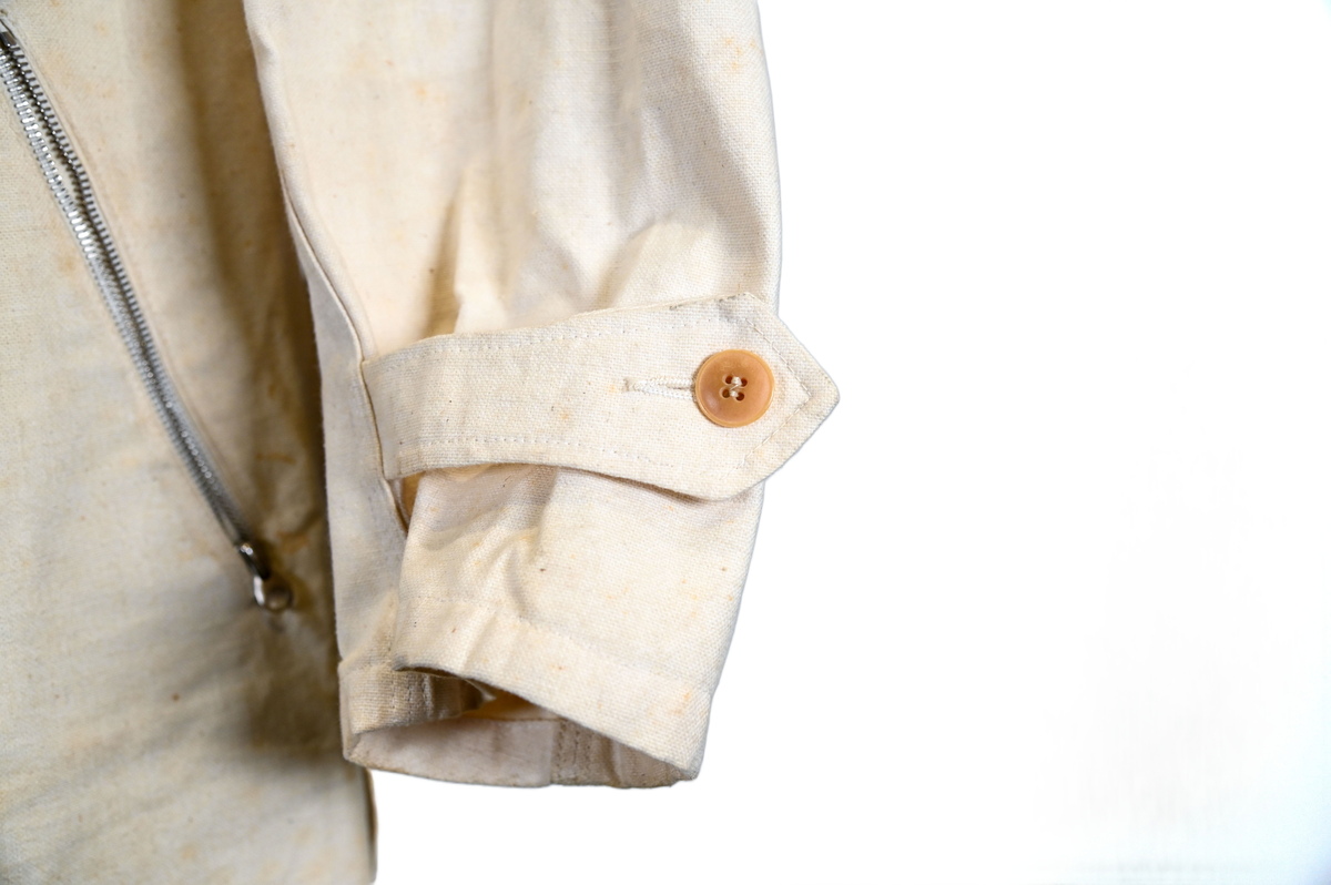 Långärmad overall i vitt tyg med krage. Overallen har två bröstfickor och två framfickor som sluts med dragkedja i metall. I midjan sitter ett bälte i samma material som plagget med ett spänne i metall. Bältet sitter på plats med med två öglor i plaggets sidor. Ärmsluten kan knäppas med tygband och knappar i plast. Bensluten kan spännas med tygband fäst i fyra öglor med metallspänne. 

Overallen är täckt av smuts och fläckar.