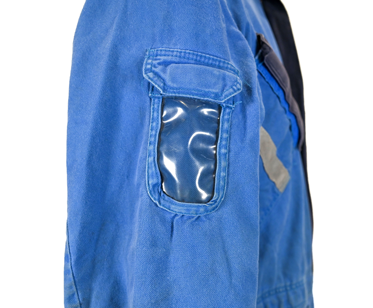 Blå arbetsoverall från SAS. På plagget finns en logga för SAS över vänster bröst och mitt på ryggen. Overallen har två bröstfickor med reflexband som försluts med kardborre. Vänster ficka saknar den del som kopplar an till kardborren på fickan. Frampå sitter två större framfickor med lock och på vardera ben sitter ytterligare två fickor på sidorna. På baksidan finns två stora bakfickor. Högt upp på sidan av vardera ärm sitter ytterligare fickor varav den högra är av genomskinlig plast. Samtliga fickor försluts med kardborre. Utöver på bröstfickorna sitter reflexband runt benens nedre del samt högt upp över ryggen. Overallen sluts med dragkedja och två tryckknappar i metall. Ärmsluten kan justeras med kardborre. Vid höfterna finns två öppningar in i overallen. På knäna finns öppningar för att lägga i knäskydd.

På plagget finns spår av vit färg och andra smutsfläckar. Vänster bakficka är nött.

Overallen är producerad av Fristads i storlek 48. På insidan längs med dragskedjan sitter en lapp med texten "OBS! DRAGKEDJAN SKALL VARA STÄNGD VID TVÄTT".