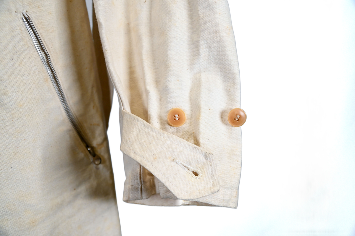 Långärmad overall i vitt tyg med krage. Overallen har två bröstfickor och två framfickor som sluts med dragkedja i metall. I midjan sitter ett bälte i samma material som plagget med ett spänne i metall. Bältet sitter på plats med med två öglor i plaggets sidor. Ärmsluten kan knäppas med tygband och knappar i plast. Bensluten kan spännas med tygband fäst i fyra öglor med metallspänne. 

Overallen är täckt av smuts och fläckar.