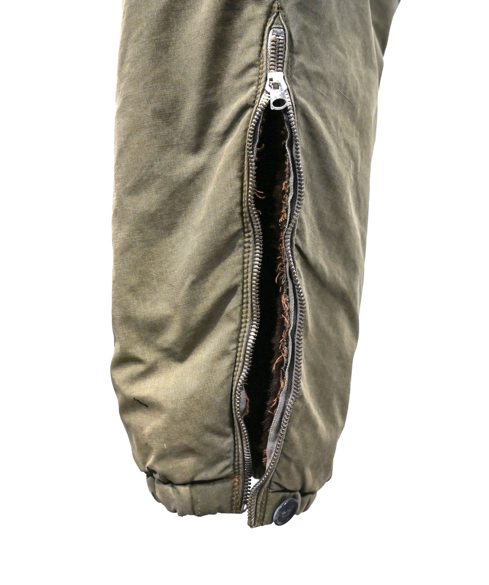 Grågrön overall som använts som flygardräkt. Liknande dräkter har använts inom flygvapnet. Dräkten är fodrad och har en fodrad krage i brun päls. I ärmsluten sitter en mörkblå resår. På framsida lår sitter en ficka på vardera ben som försluts med dragkedja i metall. På sidorna i midjan sitter en öppning som sluts med dragkedja. Dräkten stängs med en lång dragkedja i metall som sträcker sig ned i höger ben. I kragen sitter ett tygband som stänger igen kragen med en plastknapp.  Vid fotändarna finns dragkedja och knapp för att öppna upp fotsluten. 

I tygbandet på kragen satt (borttagen och lagd i ficka av vårdskäl) en lapp fasthäftad som troligtvis har fästs i plagget i samband med tvätt.