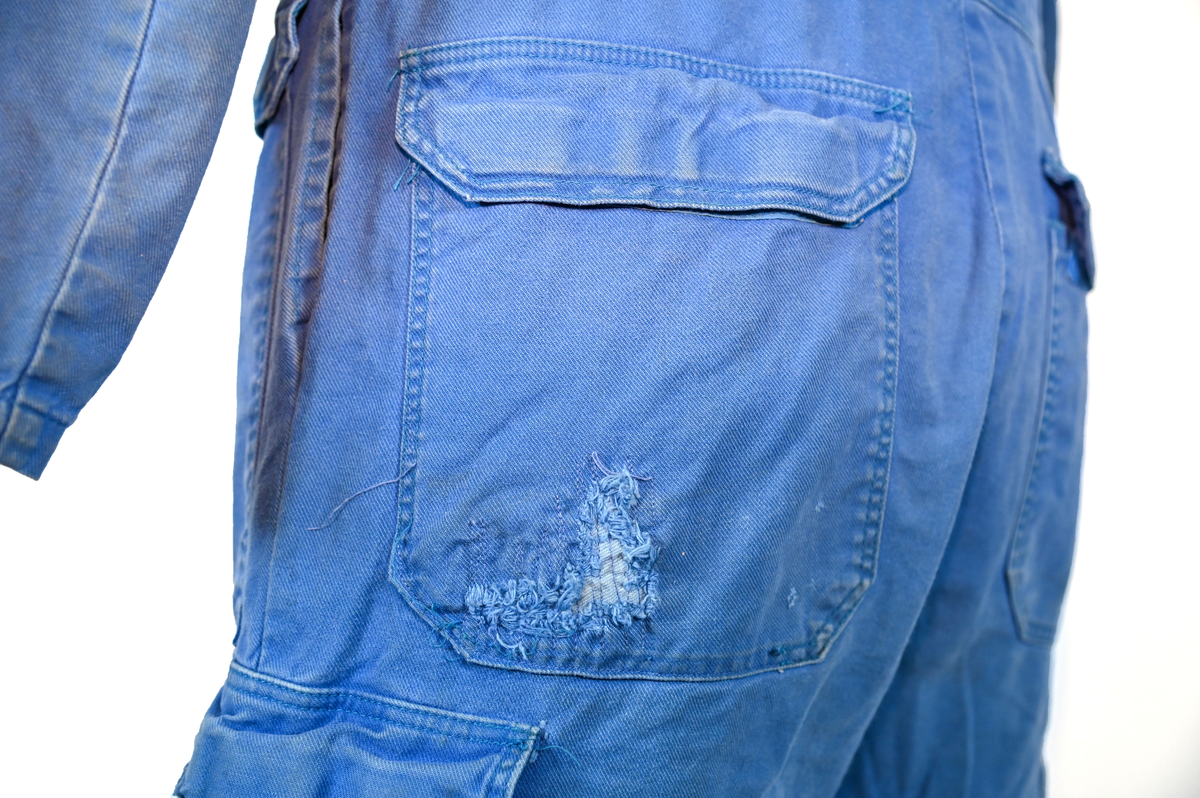 Blå arbetsoverall från SAS. På plagget finns en logga för SAS över vänster bröst och mitt på ryggen. Overallen har två bröstfickor med reflexband som försluts med kardborre. Vänster ficka saknar den del som kopplar an till kardborren på fickan. Frampå sitter två större framfickor med lock och på vardera ben sitter ytterligare två fickor på sidorna. På baksidan finns två stora bakfickor. Högt upp på sidan av vardera ärm sitter ytterligare fickor varav den högra är av genomskinlig plast. Samtliga fickor försluts med kardborre. Utöver på bröstfickorna sitter reflexband runt benens nedre del samt högt upp över ryggen. Overallen sluts med dragkedja och två tryckknappar i metall. Ärmsluten kan justeras med kardborre. Vid höfterna finns två öppningar in i overallen. På knäna finns öppningar för att lägga i knäskydd.

På plagget finns spår av vit färg och andra smutsfläckar. Vänster bakficka är nött.

Overallen är producerad av Fristads i storlek 48. På insidan längs med dragskedjan sitter en lapp med texten "OBS! DRAGKEDJAN SKALL VARA STÄNGD VID TVÄTT".