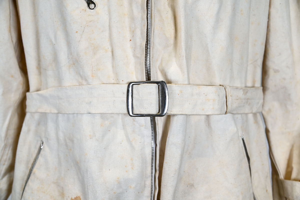 Långärmad overall i vitt tyg med krage. Overallen har två bröstfickor och två framfickor som sluts med dragkedja i metall. I midjan sitter ett bälte i samma material som plagget med ett spänne i metall. Bältet sitter på plats med med två öglor i plaggets sidor. Ärmsluten kan knäppas med tygband och knappar i plast. Bensluten kan spännas med tygband fäst i fyra öglor med metallspänne. 

Overallen är täckt av smuts och fläckar.