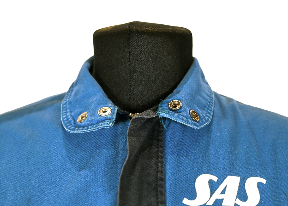 Blå arbetsoverall från SAS. På plagget finns en logga för SAS över vänster bröst och mitt på ryggen. Overallen har två bröstfickor med reflexband som försluts med kardborre. Vänster ficka saknar den del som kopplar an till kardborren på fickan. Frampå sitter två större framfickor med lock och på vardera ben sitter ytterligare två fickor på sidorna. På baksidan finns två stora bakfickor. Högt upp på sidan av vardera ärm sitter ytterligare fickor varav den högra är av genomskinlig plast. Samtliga fickor försluts med kardborre. Utöver på bröstfickorna sitter reflexband runt benens nedre del samt högt upp över ryggen. Overallen sluts med dragkedja och två tryckknappar i metall. Ärmsluten kan justeras med kardborre. Vid höfterna finns två öppningar in i overallen. På knäna finns öppningar för att lägga i knäskydd.

På plagget finns spår av vit färg och andra smutsfläckar. Vänster bakficka är nött.

Overallen är producerad av Fristads i storlek 48. På insidan längs med dragskedjan sitter en lapp med texten "OBS! DRAGKEDJAN SKALL VARA STÄNGD VID TVÄTT".