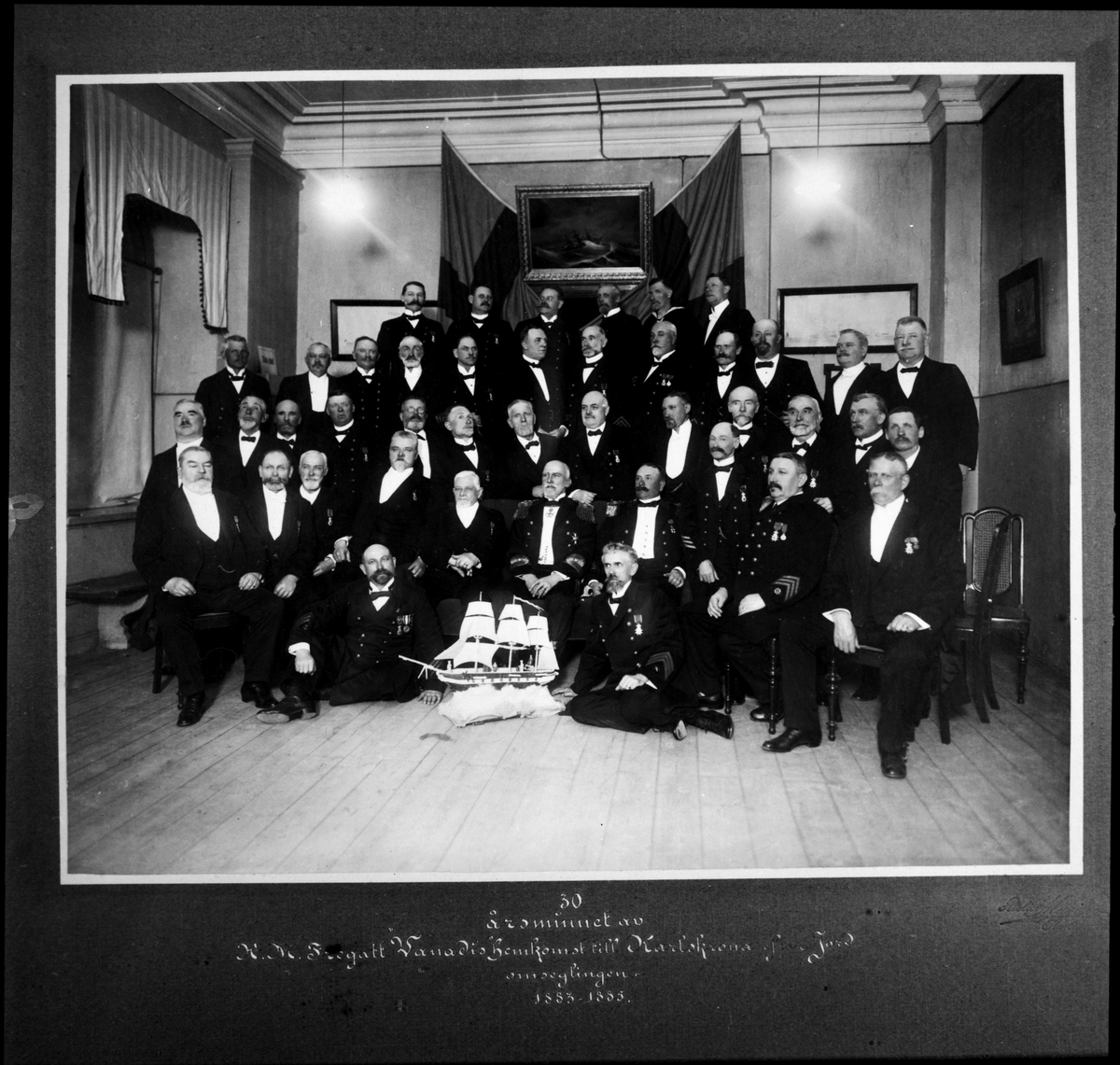 Grupporträtt av fördetta besättningsmän från fregatten Vanadis som sitter samlade bakom en modell av skeppet. Bilden togs under firandet av 30års hemkomst från världsomsegling 1883-1885. Den del är klädd i uniformer med ordenstecken på, en del bär civila kostym.