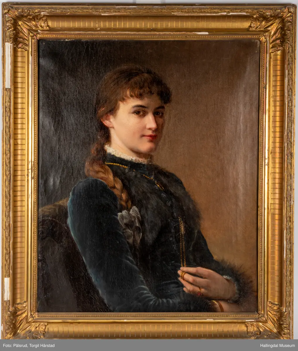 Ant. 1890-årene. Olje på lerret. Høydeformat. Kvinneportrett. Halvfigur. 3/4 prof. dreining høyre. Brun-gul bakgrunn. Sitter i en gulgrønn plysjstol med dusker, Brunt hår, krøllet pannelugg, lang flette nedover h. skulder. Blå sløyfe. Blå fløyelskjole , med høy hals og lange ermer, hvit blonde i halsen og rundt håndleddene, pelskanter på bryst og håndledd. Gullkjede med ovalt smykke rundt halsen. Venstre hånd bærer gullring med røde stener. Fingrene holder i gyllen snor med dusk, festet i kjoleåpningen. Rammen er forgylt.