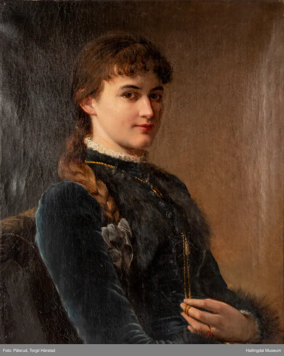 Ant. 1890-årene. Olje på lerret. Høydeformat. Kvinneportrett. Halvfigur. 3/4 prof. dreining høyre. Brun-gul bakgrunn. Sitter i en gulgrønn plysjstol med dusker, Brunt hår, krøllet pannelugg, lang flette nedover h. skulder. Blå sløyfe. Blå fløyelskjole , med høy hals og lange ermer, hvit blonde i halsen og rundt håndleddene, pelskanter på bryst og håndledd. Gullkjede med ovalt smykke rundt halsen. Venstre hånd bærer gullring med røde stener. Fingrene holder i gyllen snor med dusk, festet i kjoleåpningen. Rammen er forgylt.