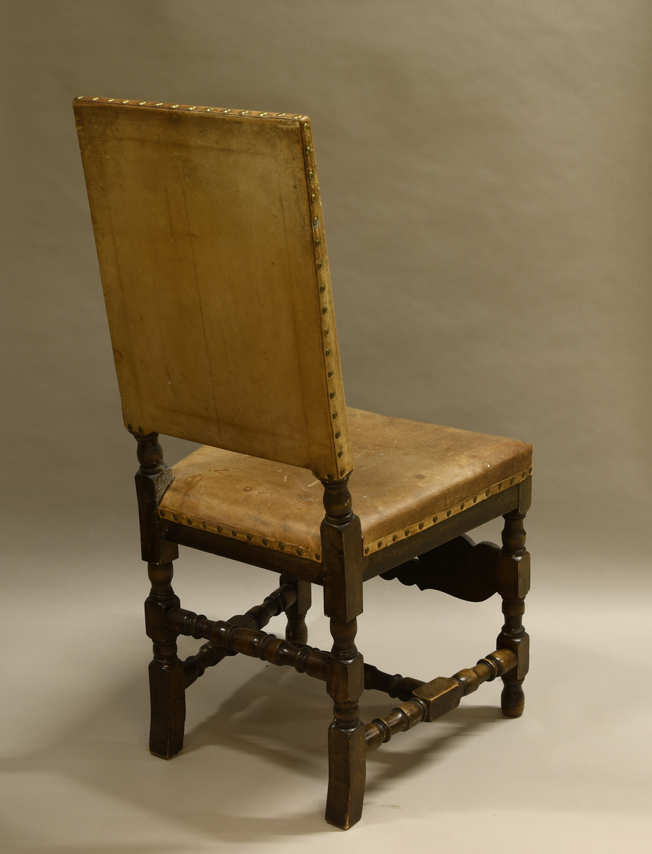 Stol av tre, med trekk av skinn. Stolen har dreide bein med rund og firkantet profil. Nederst er det en H-formet sprosse av dreid treverk, og det er en dreid sprosse mellom bakbeina. På framsiden er det en sprosse med akantusblad og i midten tre palmetter med gjennombrutte mellompartier. Ryggen har stående rektangulær form og er trukket med skinn på fram- og bakside. Setet er også trukket med skinn. Skinnet er festet med messingnagler.
