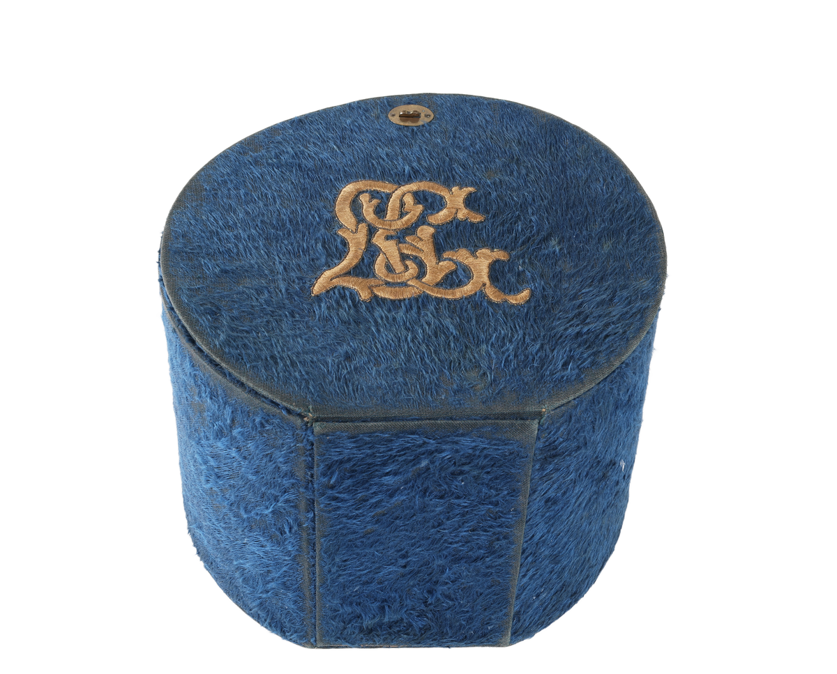 Cylindrisk ask, rak kant bak, plant lock. I framkanten lucka. Under locket cylindriskt fack för manschetter. Bakom luckan smalt cylindriskt fack för kragar. Asken klädd med plysch. På locket gulbrunt monogram i plattsöm "LC". Inuti klädd med vitt papper. Ett knäppe saknas. Bruksslitage.

Funktion: Förvaring av kragar och manschetter