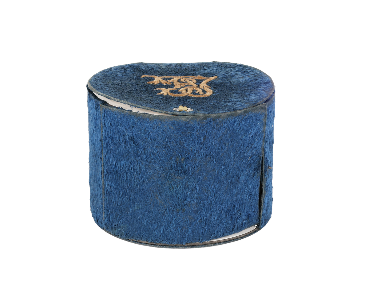 Cylindrisk ask, rak kant bak, plant lock. I framkanten lucka. Under locket cylindriskt fack för manschetter. Bakom luckan smalt cylindriskt fack för kragar. Asken klädd med plysch. På locket gulbrunt monogram i plattsöm "LC". Inuti klädd med vitt papper. Ett knäppe saknas. Bruksslitage.

Funktion: Förvaring av kragar och manschetter