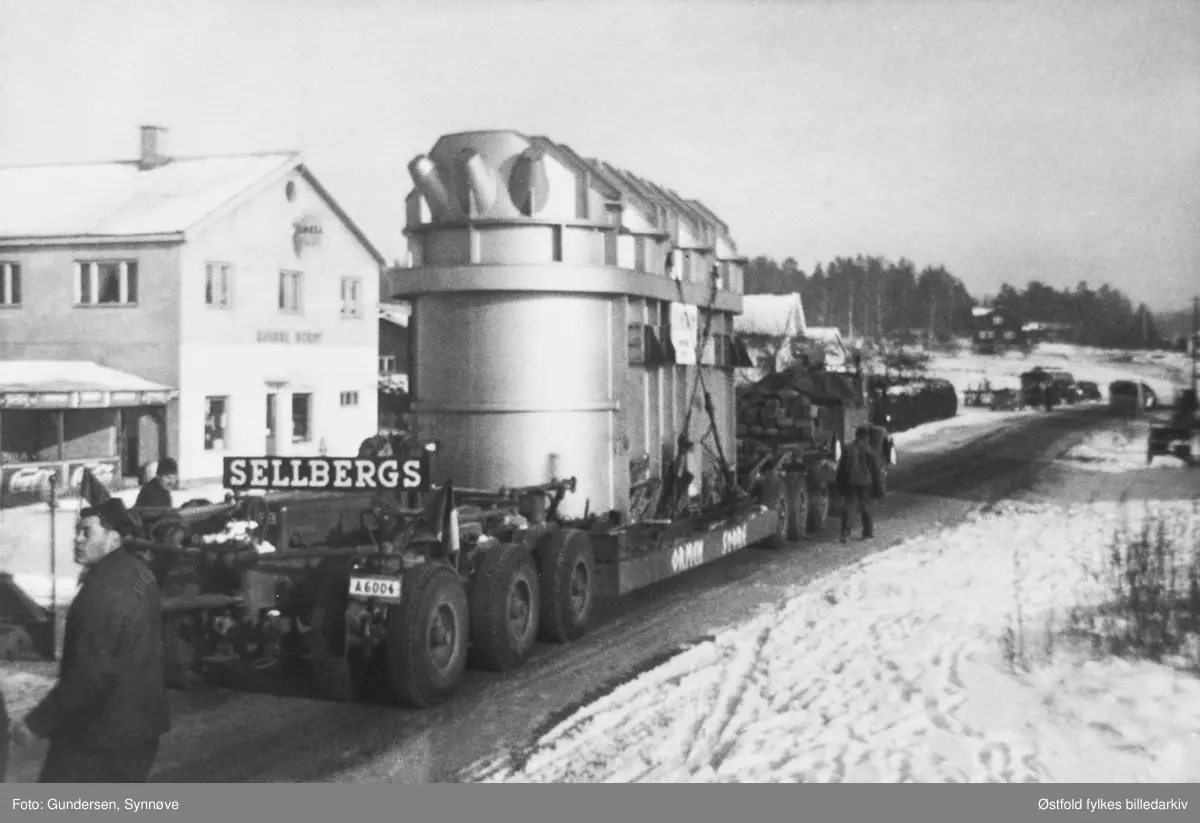 Transport av transformator fra Ise stasjon til Hasle i Varteig juli 1963. Bildet er tatt like ved Bj. Berbys bensinstasjon på Ise. Boggien (flerakslet bakstell på lastebiler og tilhengere (trailere)) veide 46 tonn. På siden av boggien er det malt "ORMEN STORE", firmamerke "Sellbergs". Trafoen veide 140 tonn. Bilkjennetegn: A-6004.