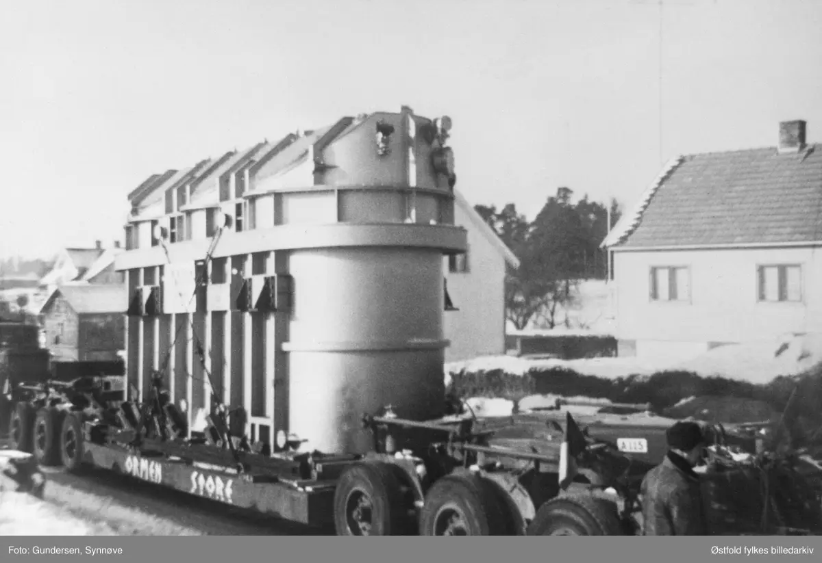 Transport av transformator fra Ise stasjon til Hasle, Varteig i juli 1963. Bildet er tatt like ved Bj. Berbys bensinstasjon på Ise. Boggien (flerakslet bakstell på lastebiler og tilhengere (trailere)) veide 46 tonn. På siden av boggien er det malt "ORMEN STORE", firmamerke "Sellbergs". Trafoen veide 140 tonn.