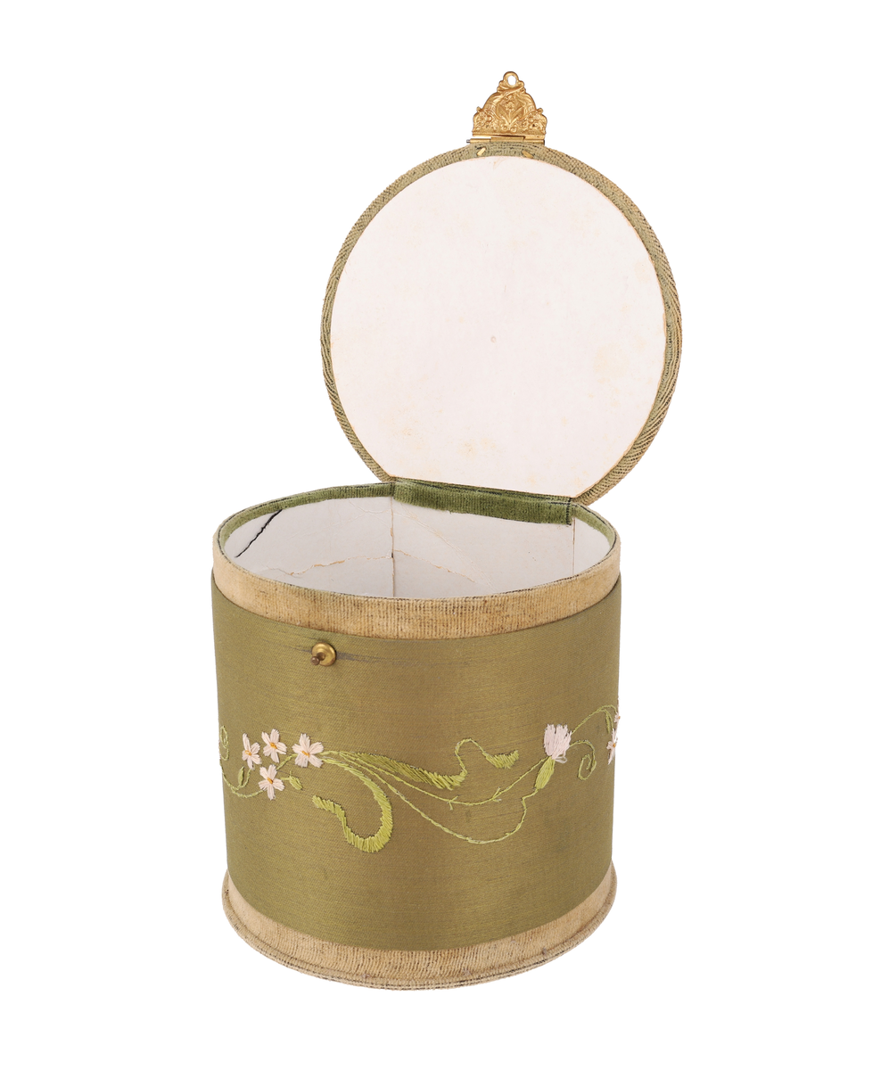 Närmast cylindrisk ask med plant lock. Klädd med grön bomullssammet, brett band av atlassiden med broderad blomslinga i silketråd. Handtag och lås av förgyllt metallbleck. Stomme i vit papp. I gott skick.