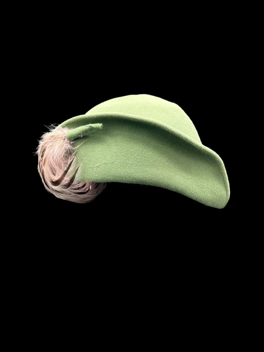 Grön damhatt med fjädrar och hattnål. Kulle och brätte formad av ett stycke styvt tyg. Brättet går inte hela vägen runt och är bara delvis uppvikt. På framsidan av den uppvikta delen av brättet finns ett knippe fjädrar sammanhållna med ett stycke av samma material som i hatten. På insidan ett grått ripsband. Ofodrad. En hattnål av metall vars huvud är överklätt med samma tyg som i hatten. Ett flätat band runt kullen av remsor av samma material som i övriga hatten. Elastiskt brunt band från ena sidan av hatten till den andra för fastsättning.