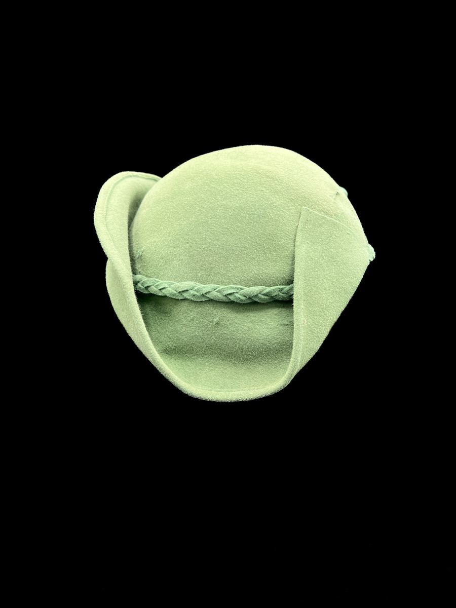 Grön damhatt med fjädrar och hattnål. Kulle och brätte formad av ett stycke styvt tyg. Brättet går inte hela vägen runt och är bara delvis uppvikt. På framsidan av den uppvikta delen av brättet finns ett knippe fjädrar sammanhållna med ett stycke av samma material som i hatten. På insidan ett grått ripsband. Ofodrad. En hattnål av metall vars huvud är överklätt med samma tyg som i hatten. Ett flätat band runt kullen av remsor av samma material som i övriga hatten. Elastiskt brunt band från ena sidan av hatten till den andra för fastsättning.