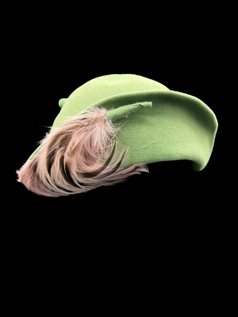 Grön damhatt med fjädrar och hattnål. Kulle och brätte formad av ett stycke styvt tyg. Brättet går inte hela vägen runt och är bara delvis uppvikt. På framsidan av den uppvikta delen av brättet finns ett knippe fjädrar sammanhållna med ett stycke av samma material som i hatten. På insidan ett grått ripsband. Ofodrad. En hattnål av metall vars huvud är överklätt med samma tyg som i hatten. Ett flätat band runt kullen av remsor av samma material som i övriga hatten. Elastiskt brunt band från ena sidan av hatten till den andra för fastsättning.