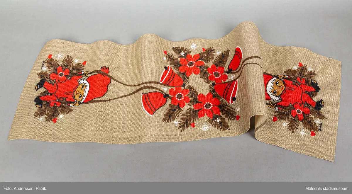 En avlång jullöpare i färgerna natur, röd och vit. På löparens mitt finns julklockor och röda julstjärnor. En rödklädd tomte på löparens ände håller i band som sammanbinds med klockorna. Tomten har röd tomtemössa.