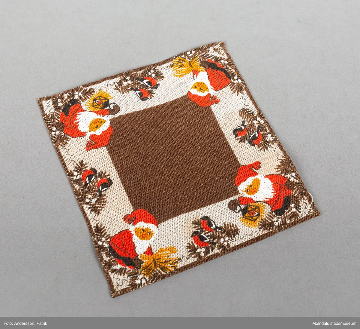 En liten kvadratisk julduk. Ytterkanten är beige och dekorerad med en rödklädd tomte i varje hörn. Mellan tomtarna finns ett grankvistmotiv med talgoxar. Tomtarna håller i en lykta.
Mönstet är symmetriskt i dukens ytterkant.
Juldukarna har tillhört Elisabeth Åberg som är "juloman" och älskar julen.