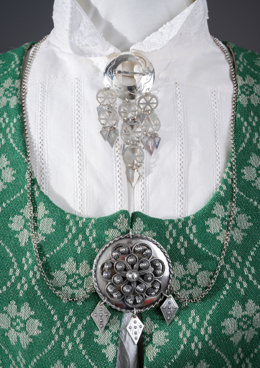 Trønderbunad med grønn ullstakk. Grønn vest med mønster "rose med bølge", ulldamask med hvit lin i renning og ull i innslag. Veske i sort ullstoff med flerfarget broderi med mønster "sokndal rust", bakstykke av skinn, foret med baskerlin, forsølvet veskelås. Skjorte av hvit lin, rikt broderte mansjetter, halslinning og skulderstykke, såkalt "festskjorte" med rankesøm og uttrekkssøm på ermene og brystet.