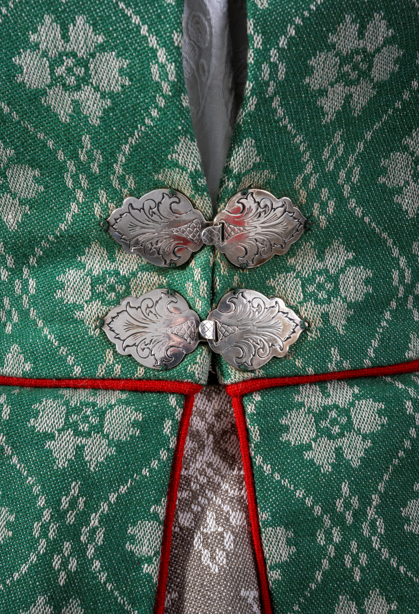 Trønderbunad med grønn ullstakk. Grønn vest med mønster "rose med bølge", ulldamask med hvit lin i renning og ull i innslag. Veske i sort ullstoff med flerfarget broderi med mønster "sokndal rust", bakstykke av skinn, foret med baskerlin, forsølvet veskelås. Skjorte av hvit lin, rikt broderte mansjetter, halslinning og skulderstykke, såkalt "festskjorte" med rankesøm og uttrekkssøm på ermene og brystet.
