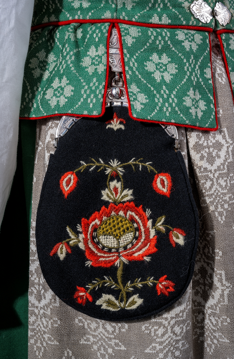 Trønderbunad med grønn ullstakk. Grønn vest med mønster "rose med bølge", ulldamask med hvit lin i renning og ull i innslag. Veske i sort ullstoff med flerfarget broderi med mønster "sokndal rust", bakstykke av skinn, foret med baskerlin, forsølvet veskelås. Skjorte av hvit lin, rikt broderte mansjetter, halslinning og skulderstykke, såkalt "festskjorte" med rankesøm og uttrekkssøm på ermene og brystet.