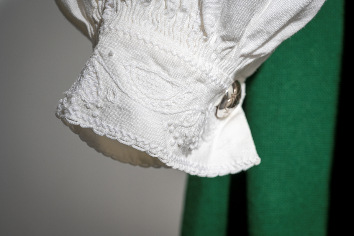Trønderbunad med grønn ullstakk. Grønn vest med mønster "rose med bølge", ulldamask med hvit lin i renning og ull i innslag. Veske i sort ullstoff med flerfarget broderi med mønster "sokndal rust", bakstykke av skinn, foret med baskerlin, forsølvet veskelås. Skjorte av hvit lin, rikt broderte mansjetter, halslinning og skulderstykke, såkalt "festskjorte" med rankesøm og uttrekkssøm på ermene og brystet.