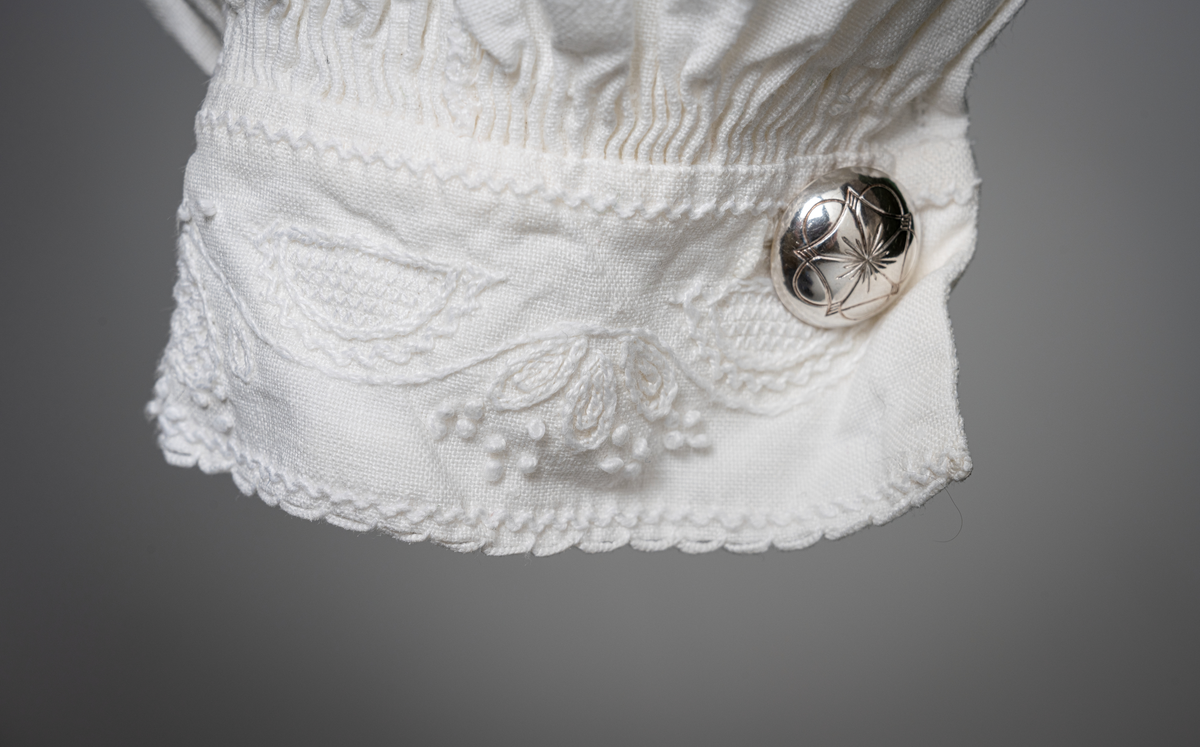 Trønderbunad med grønn ullstakk. Grønn vest med mønster "rose med bølge", ulldamask med hvit lin i renning og ull i innslag. Veske i sort ullstoff med flerfarget broderi med mønster "sokndal rust", bakstykke av skinn, foret med baskerlin, forsølvet veskelås. Skjorte av hvit lin, rikt broderte mansjetter, halslinning og skulderstykke, såkalt "festskjorte" med rankesøm og uttrekkssøm på ermene og brystet.