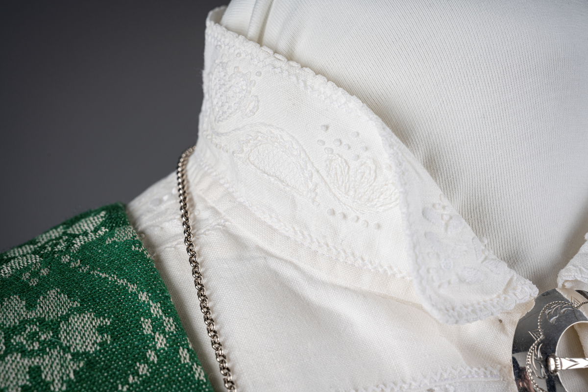 Trønderbunad med grønn ullstakk. Grønn vest med mønster "rose med bølge", ulldamask med hvit lin i renning og ull i innslag. Veske i sort ullstoff med flerfarget broderi med mønster "sokndal rust", bakstykke av skinn, foret med baskerlin, forsølvet veskelås. Skjorte av hvit lin, rikt broderte mansjetter, halslinning og skulderstykke, såkalt "festskjorte" med rankesøm og uttrekkssøm på ermene og brystet.