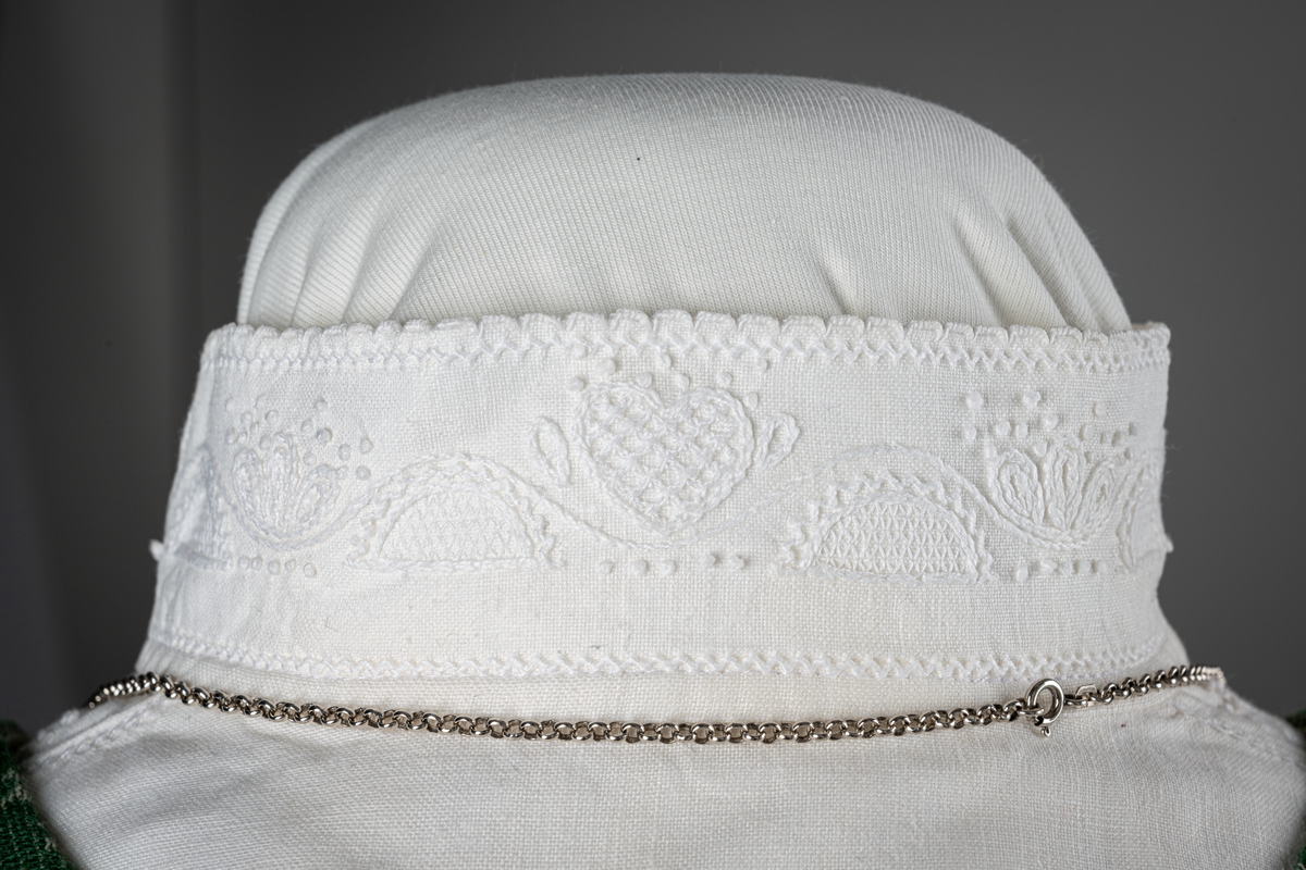 Trønderbunad med grønn ullstakk. Grønn vest med mønster "rose med bølge", ulldamask med hvit lin i renning og ull i innslag. Veske i sort ullstoff med flerfarget broderi med mønster "sokndal rust", bakstykke av skinn, foret med baskerlin, forsølvet veskelås. Skjorte av hvit lin, rikt broderte mansjetter, halslinning og skulderstykke, såkalt "festskjorte" med rankesøm og uttrekkssøm på ermene og brystet.