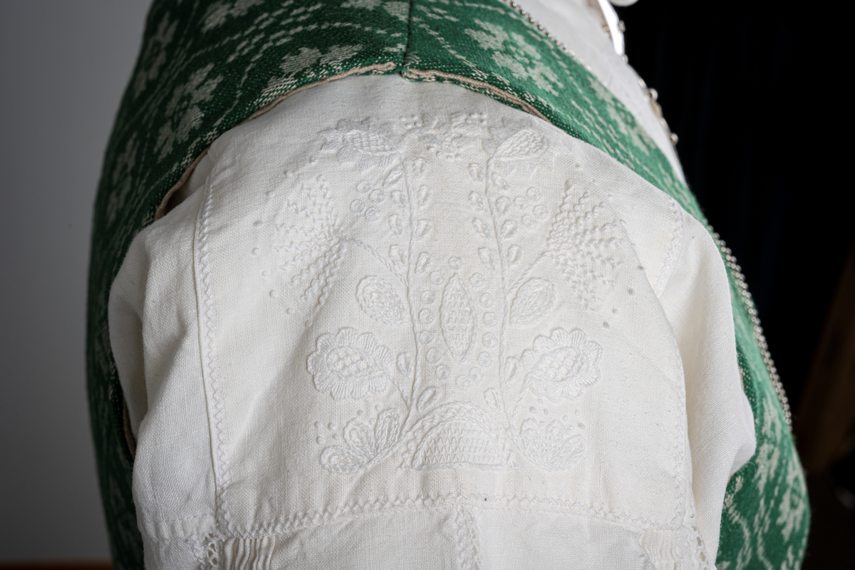 Trønderbunad med grønn ullstakk. Grønn vest med mønster "rose med bølge", ulldamask med hvit lin i renning og ull i innslag. Veske i sort ullstoff med flerfarget broderi med mønster "sokndal rust", bakstykke av skinn, foret med baskerlin, forsølvet veskelås. Skjorte av hvit lin, rikt broderte mansjetter, halslinning og skulderstykke, såkalt "festskjorte" med rankesøm og uttrekkssøm på ermene og brystet.