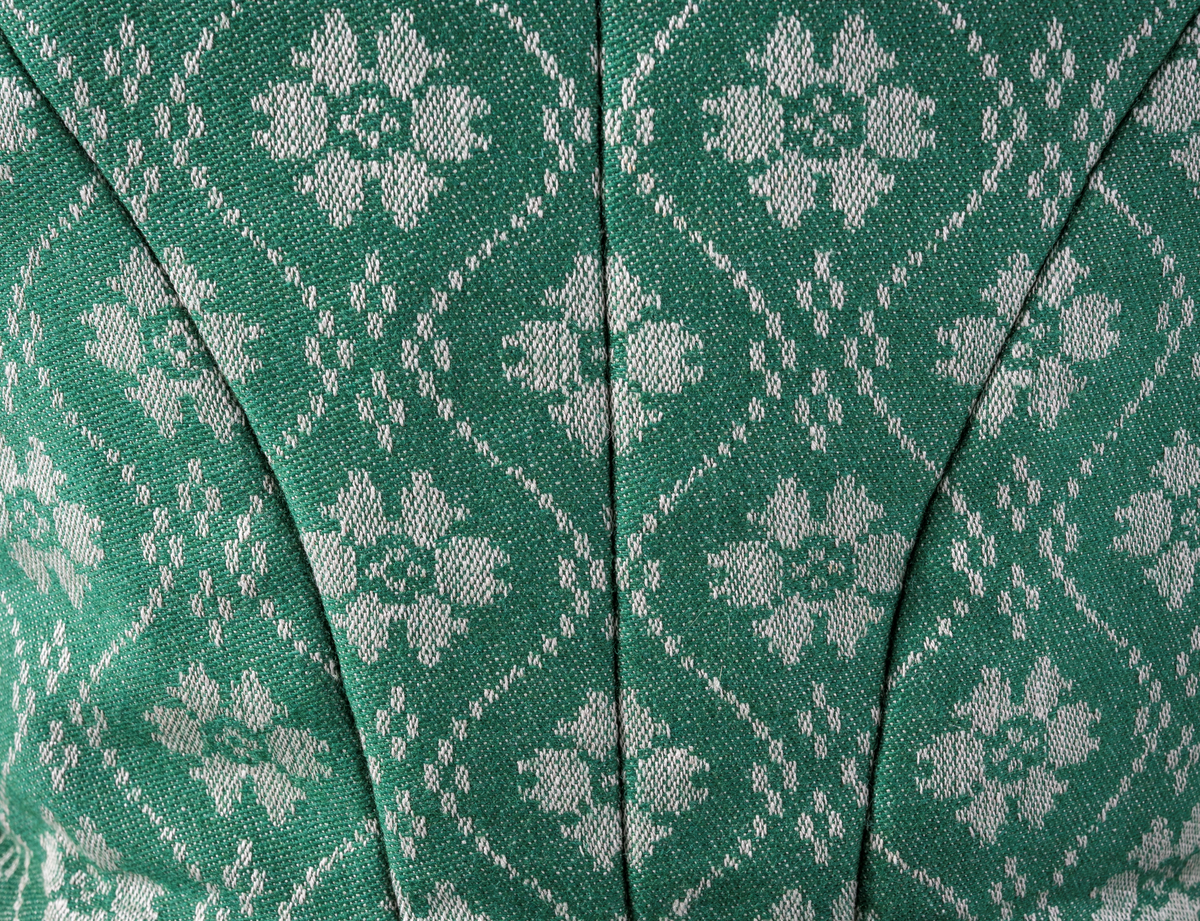 Trønderbunad med grønn ullstakk. Grønn vest med mønster "rose med bølge", ulldamask med hvit lin i renning og ull i innslag. Veske i sort ullstoff med flerfarget broderi med mønster "sokndal rust", bakstykke av skinn, foret med baskerlin, forsølvet veskelås. Skjorte av hvit lin, rikt broderte mansjetter, halslinning og skulderstykke, såkalt "festskjorte" med rankesøm og uttrekkssøm på ermene og brystet.
