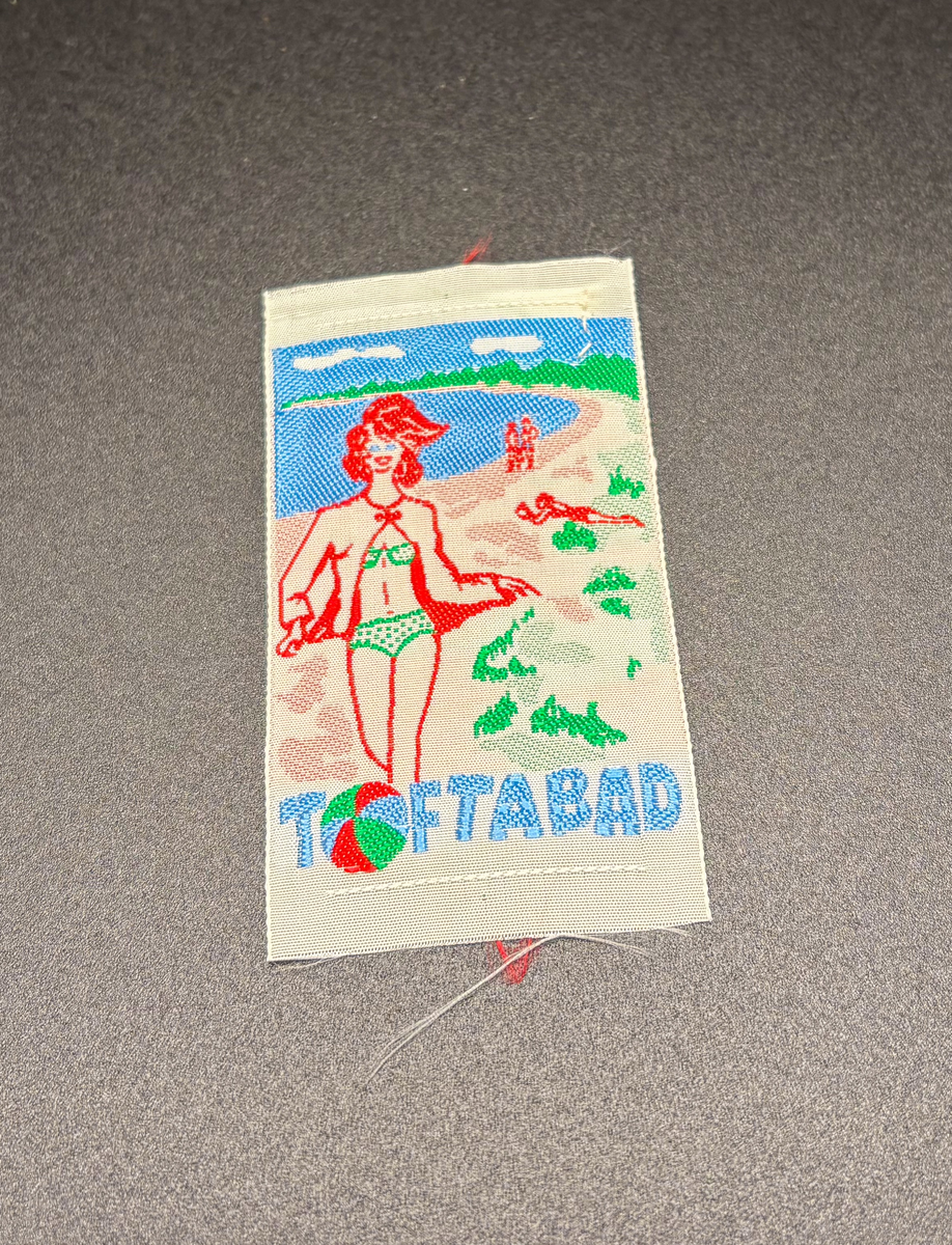 Souvenir, tygmärken att sy på kläder / väska, 3 st med olika gotlandsmotiv. Märke 1: Gotlands vapen, murgröna samt texten "GOTLAND" mot svart botten. Märke 2: Visbys vapen, del av muren samt texten "VISBY" i rött och gult mot vit botten. Märke 3: Strand med solbadande människor, i förgrunden en bikiniklädd kvinna samt texten "TOFTABAD" med en badboll som "O". Del av samling souvenirer som kommit i donatorns ägo genom en släkting. En del av materialet kan även vara insamlat av Gotlands Konstmuseum inför en utställning (dock inte troligt).