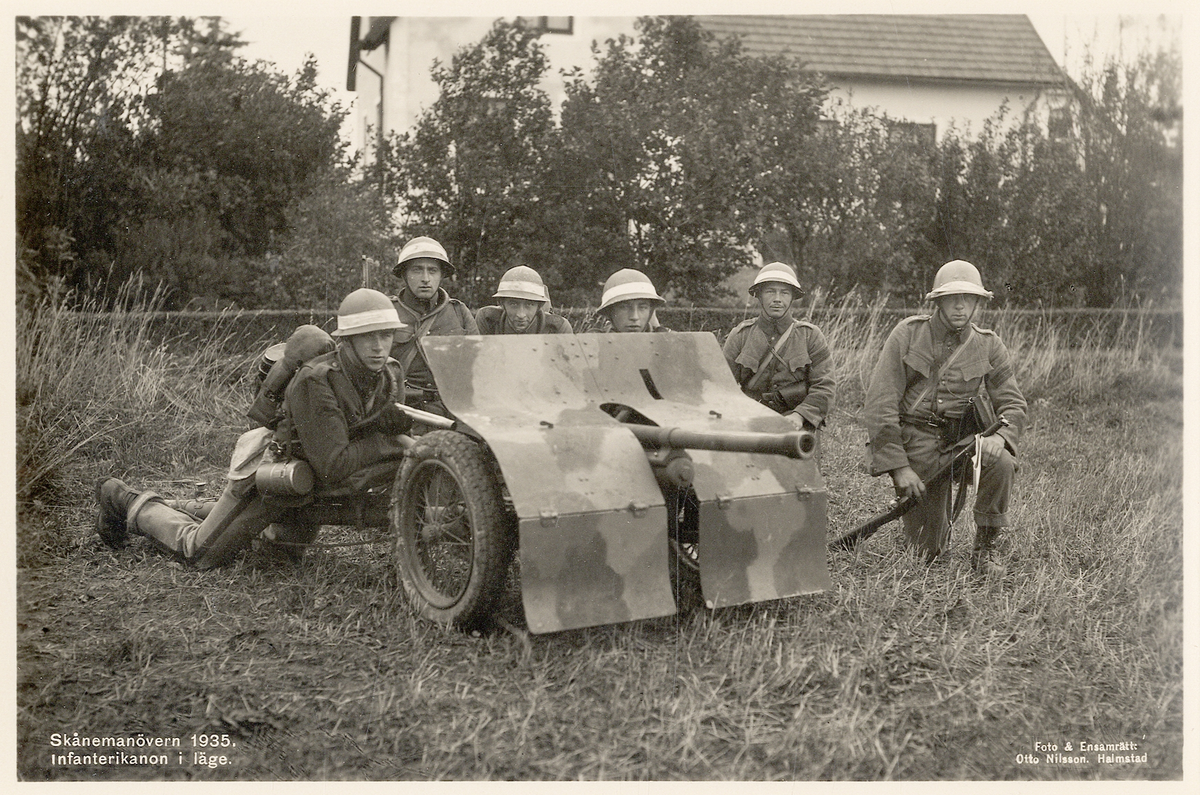 Skånemanövern 1935. 
En kamouflerad infanterikanon i läge, skött av sex väl förberedda mannar. I bakgrunden skymtar ett bostadshus.