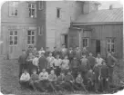 Män, ungdomar och pojkar uppställda utanför en gelbgjutarverkstad i Gränna. Anställda vid fabrikör A E Bollings gelbgjutareverkstad. Två kvinnor tittar ut genom ett fönster på andra våningen.