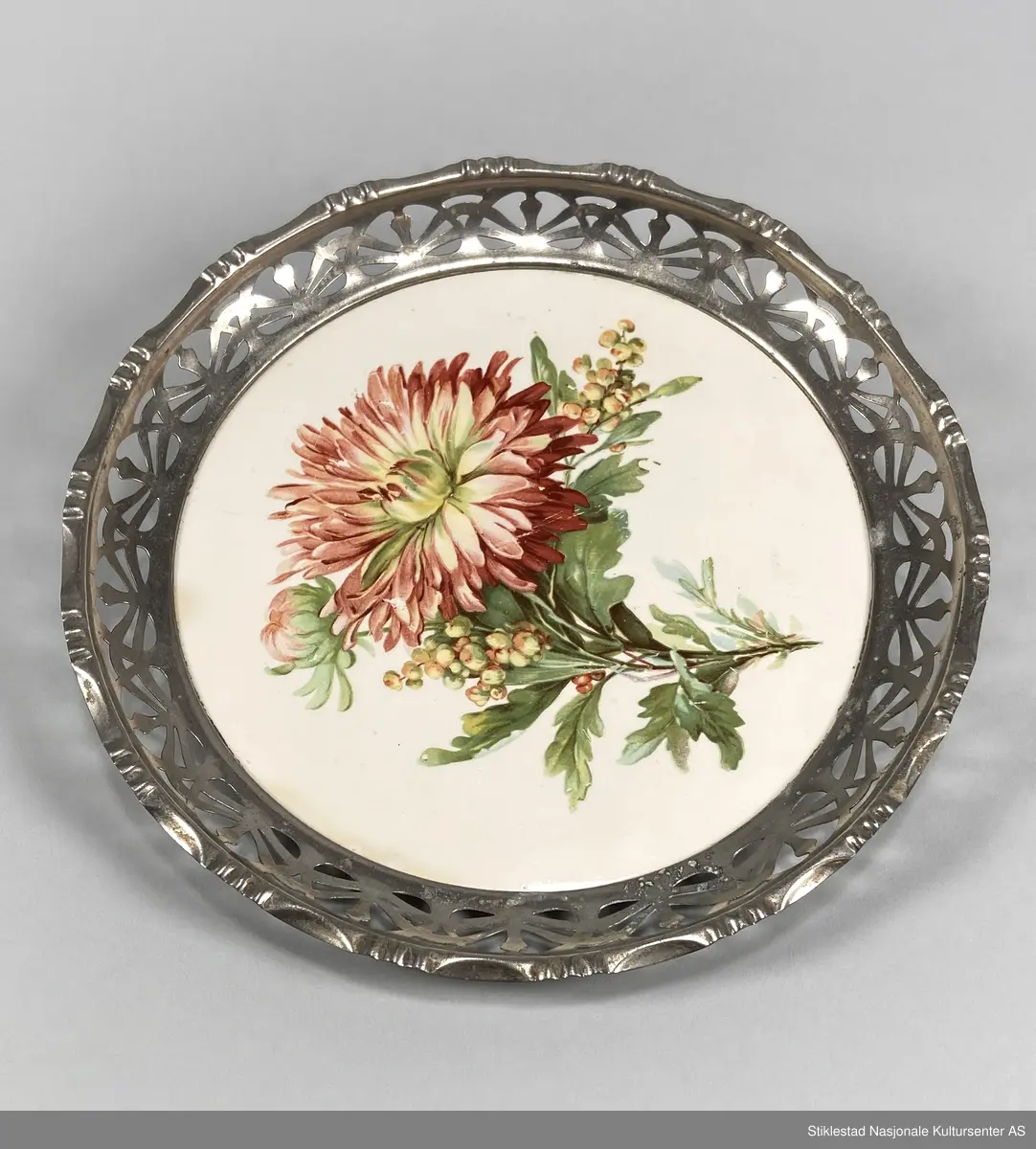 Fabrikkprodusert, rundt serveringsbrett/brødfat med gjenombrutt mønster i metallkant og bunn av keramikkflis? 
Blomster med bladverk påtrykt bunnen (antatt gerorgine/dahlia). 
Rammen har skråstilte, oppstående sider. Tysk produksjon