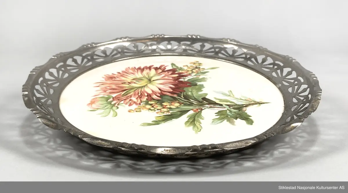 Fabrikkprodusert, rundt serveringsbrett/brødfat med gjenombrutt mønster i metallkant og bunn av keramikkflis? 
Blomster med bladverk påtrykt bunnen (antatt gerorgine/dahlia). 
Rammen har skråstilte, oppstående sider. Tysk produksjon