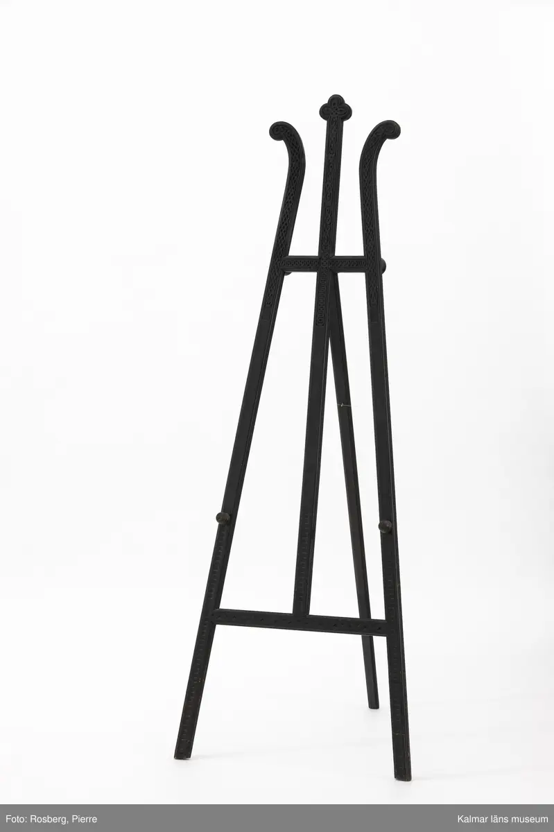 KLM 28494:130. Staffli. Tillverkat av trä, tre ben, svartmålat med utskuren ornamentik. Två rundsvarvade knoppar, stöd till tavla. Fiskelina dragen genom tre skruvöglor som stöd mellan benen.