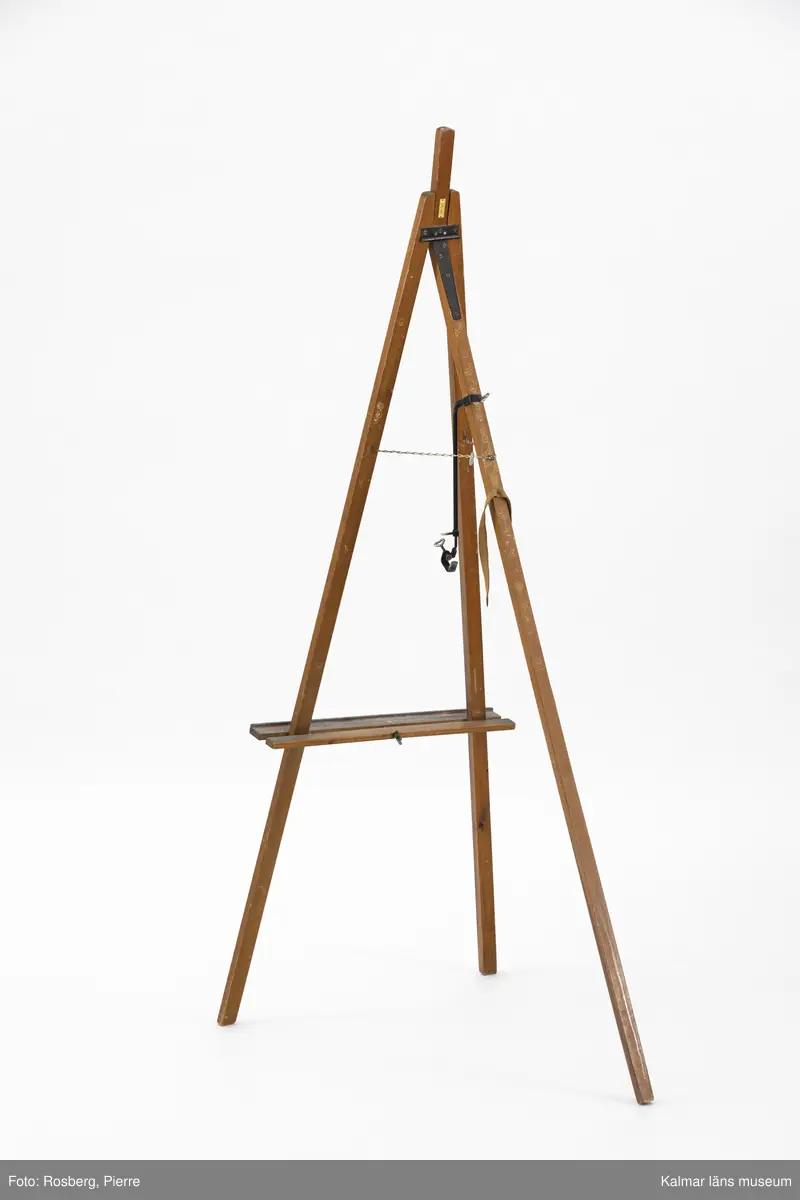 KLM 46734:1. Staffli. Golvstaffli av trä med tre ben, kedja och skruvstöd av metall. Rem av textil  på bakre benet, används i hopfällt läge. På baksidan tillverkarmärke av mässing med text: Beckers MADE IN SWEDEN. I kedjan hänger ett runt företagsmärke av metall och papper med text: A.B. WILH. BECKER KONSTNÄRSMATERIAL. Löst skruvstöd, del till staffli.