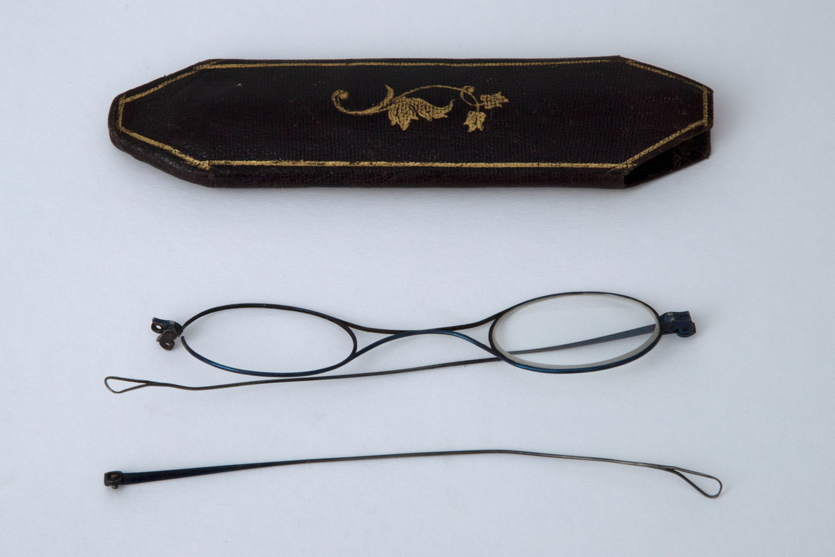 Briller i etui. Små ovale glass i innfatning av tynn stålstreng med hengslede, rette stenger som er svakt bøyd og har hempeform ved avslutningen. Etuiet er smalt med åpning i den ene enden. Trukket med tynt, brunt skinn med innpressede gullornamenter.