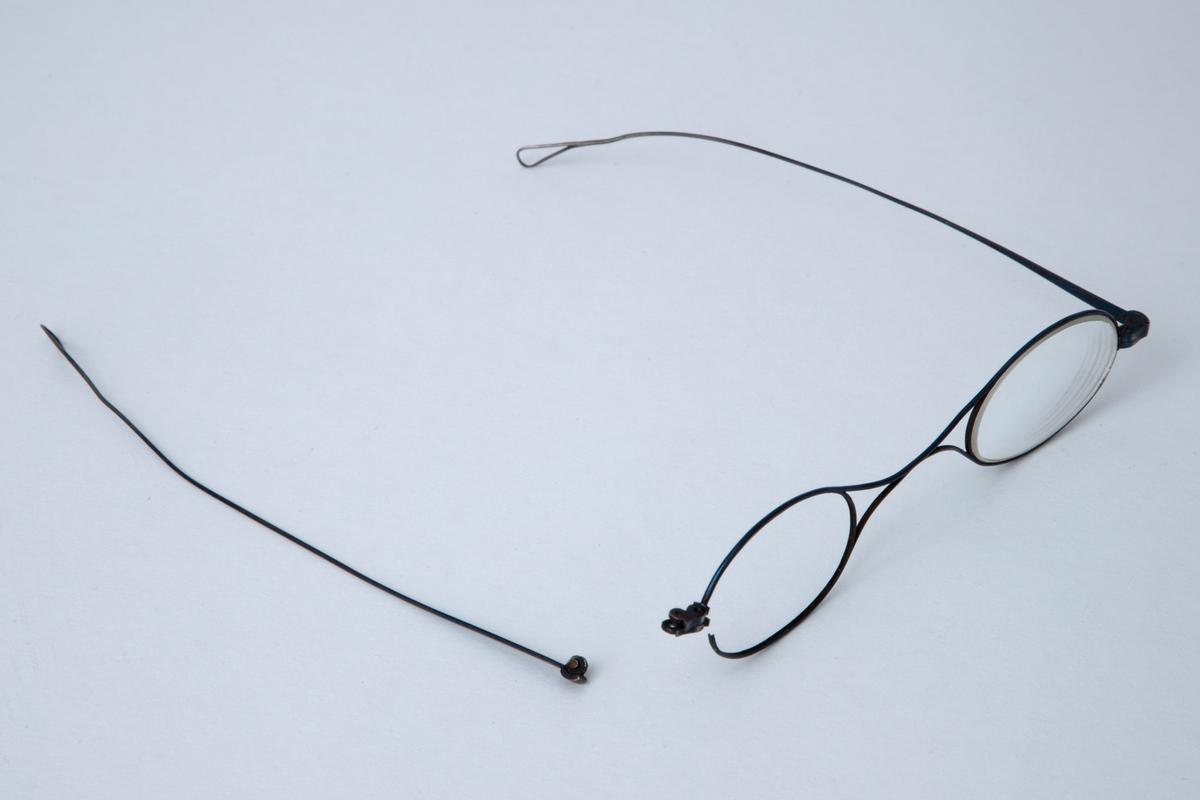 Briller i etui. Små ovale glass i innfatning av tynn stålstreng med hengslede, rette stenger som er svakt bøyd og har hempeform ved avslutningen. Etuiet er smalt med åpning i den ene enden. Trukket med tynt, brunt skinn med innpressede gullornamenter.