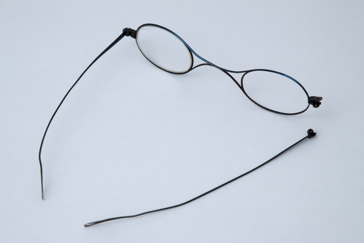 Briller i etui. Små ovale glass i innfatning av tynn stålstreng med hengslede, rette stenger som er svakt bøyd og har hempeform ved avslutningen. Etuiet er smalt med åpning i den ene enden. Trukket med tynt, brunt skinn med innpressede gullornamenter.