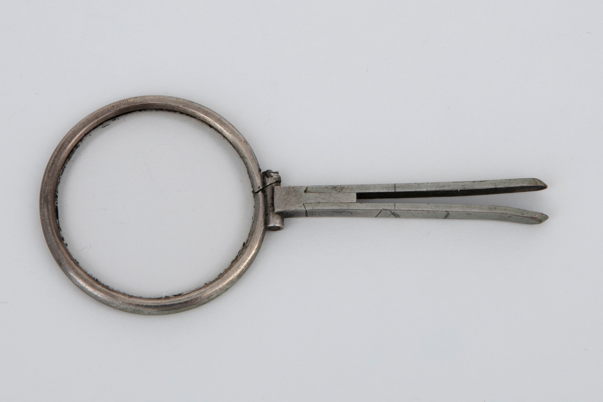 Seglass i etui. Sirkelrund glass i metallinnfatning, antagelig sølv. Til rammen festet et kort gaffelformet skaft av samme metall. Oval etui av stiv papp med fast lokk, kledd med rødt skinn og fóret med rød fløyel. På lokket blomsterornamenter i gulltrykk og initialerne: F. K.