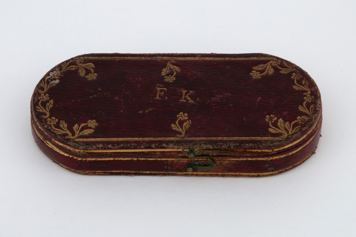 Seglass i etui. Sirkelrund glass i metallinnfatning, antagelig sølv. Til rammen festet et kort gaffelformet skaft av samme metall. Oval etui av stiv papp med fast lokk, kledd med rødt skinn og fóret med rød fløyel. På lokket blomsterornamenter i gulltrykk og initialerne: F. K.