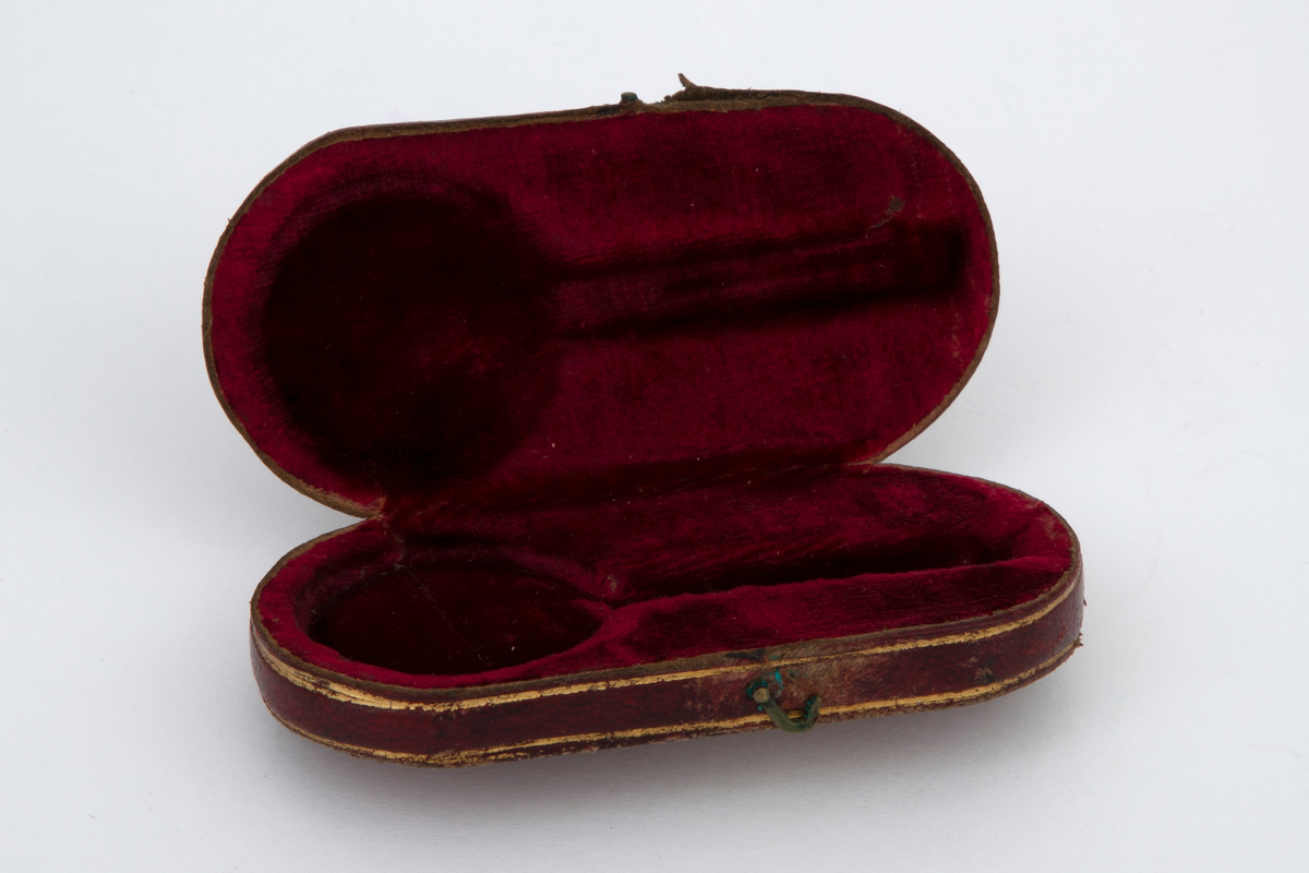 Seglass i etui. Sirkelrund glass i metallinnfatning, antagelig sølv. Til rammen festet et kort gaffelformet skaft av samme metall. Oval etui av stiv papp med fast lokk, kledd med rødt skinn og fóret med rød fløyel. På lokket blomsterornamenter i gulltrykk og initialerne: F. K.