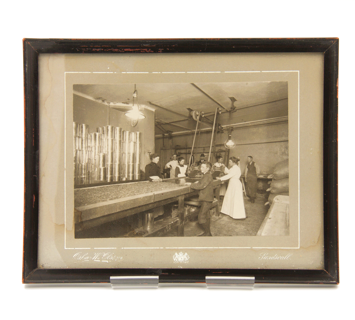 Inramad fotografi av Oskar Wilhelm Olsson (1870-1922), Sundsvall, med en fabriksinteriör som huvudmotiv. Anställda är involverade i konfektyrproduktion (?), omgivna av plåtburkar. Mörkröd träram och glas. Omfattande fuktskador på baksidan."