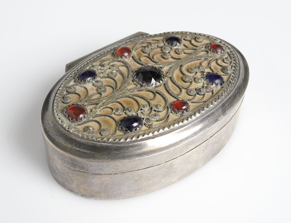 Dosa av silver, med lockOval modell, invändigt förgylld. Locket är försett med filigransarbete som har infattade blå och röda glasflusser. Stämplar i bottnens utsida. Stadsstämpel saknas.

Inskrivet i huvudbok 1988.