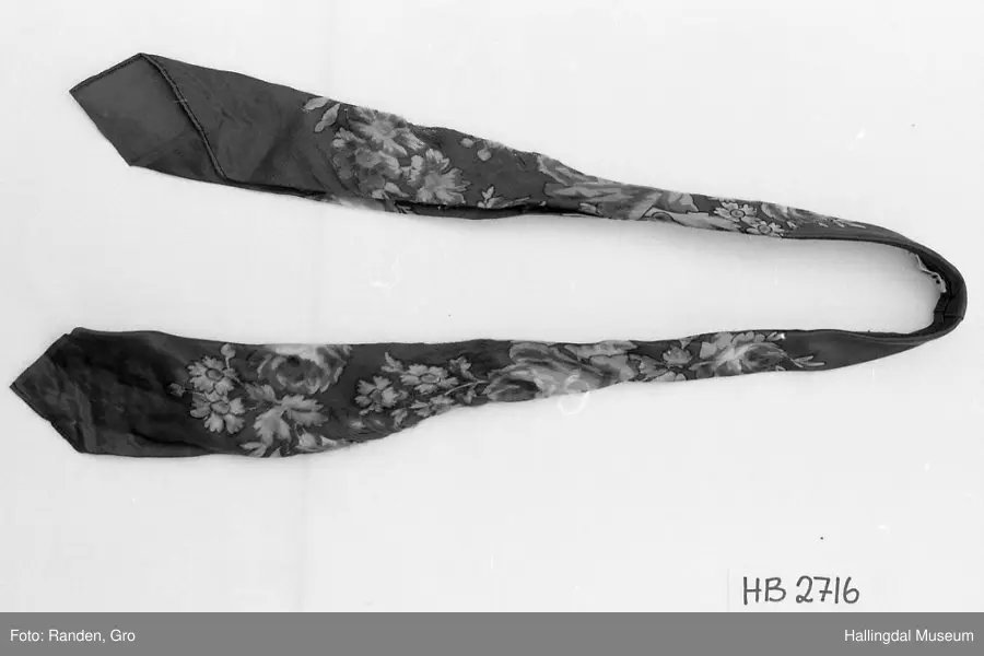 Halseklut "bastslør". Rød silke med røde, gule, grønne og blå blomster. Brettet og sydd sammen til et slips. L: 98 cm, bredde: 3,5 cm.