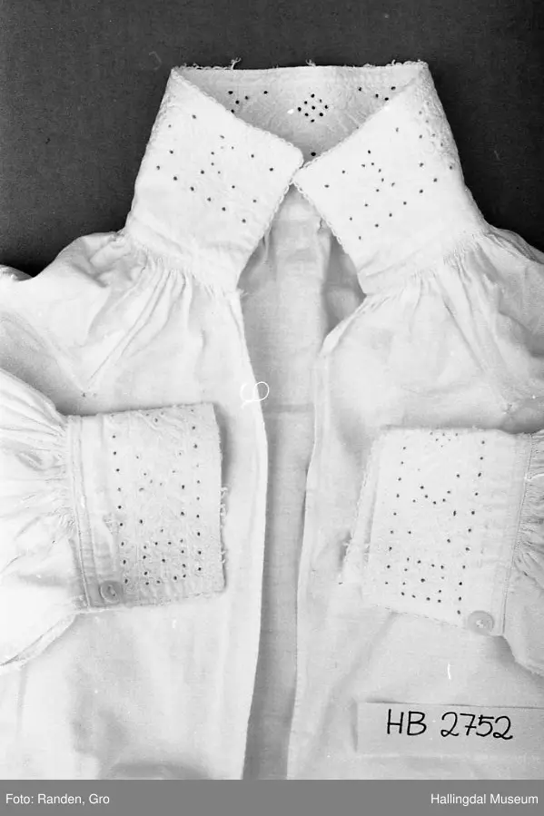 Kvit skjorte med opning framme. Hals- og ermelinningar er brodert i kvitsaum. 