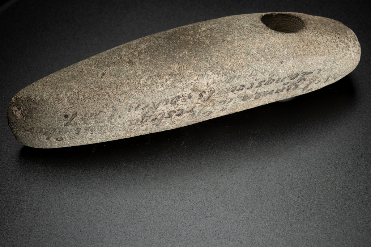 Skafthålsyxa av en ljus, relativt finkornig bergart, möjligen grönsten. Lång och ovanligt smal. Skafthålet placerat långt bakåt. Slipad.