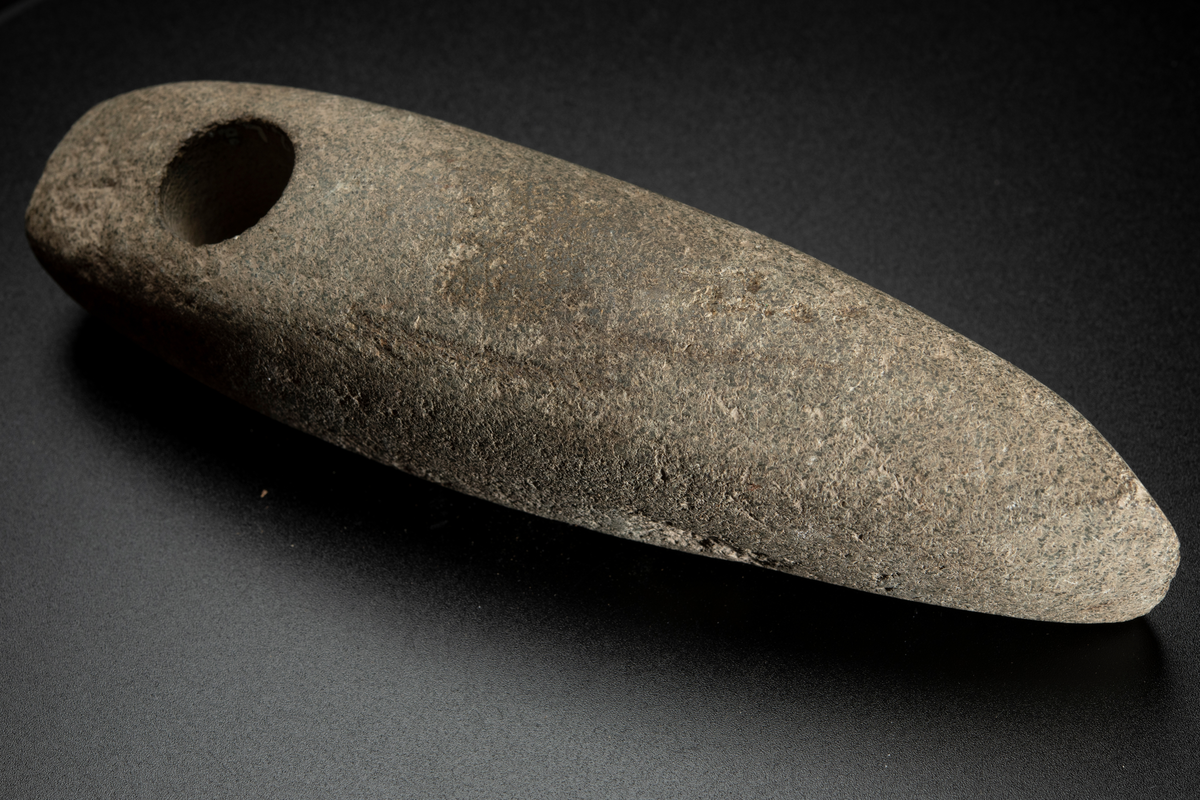 Skafthålsyxa av en ljus, relativt finkornig bergart, möjligen grönsten. Lång och ovanligt smal. Skafthålet placerat långt bakåt. Slipad.