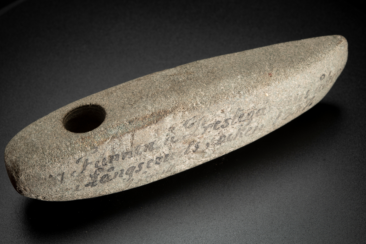 Skafthålsyxa av en ljus, relativt finkornig bergart, möjligen grönsten. Lång och ovanligt smal. Skafthålet placerat långt bakåt. Slipad.